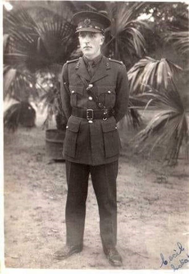 A young man, Cecil Kilpatrick, in a uniform stands for a photograph. In the background are palm fronds.