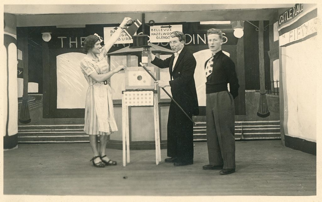 A photograph of prisoners of war in an arranged play. Two men stand in fancy clothes to the left of a magic trick, while one man in a dress stands to the left. In the background, signs point to the right reading: Bellevue, Hazelwood, and Glengo-, with the last word obscured. A sign points left and appears to read: Finaghy, Lisburn.  