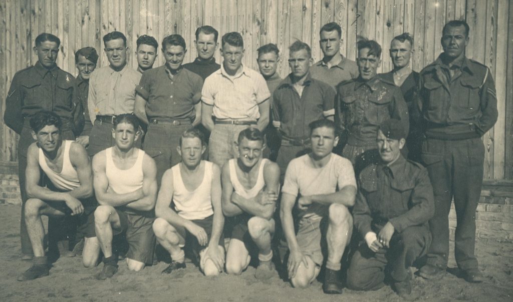 19 British servicemen, arranged in three rows, pose for a photograph in their prisoner of war camp. They are in varying states of uniform - some in full with their hat on, some in a shirt and trousers, some in an undershirt and shorts.