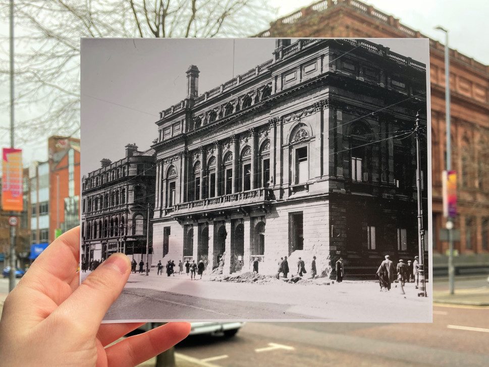 An archival photograph of Belfast Central Library, taken in the aftermath of the Blitz, is held up in front of the Library as it is today. The archival photograph shows some damage in front of the library caused by the bombing.