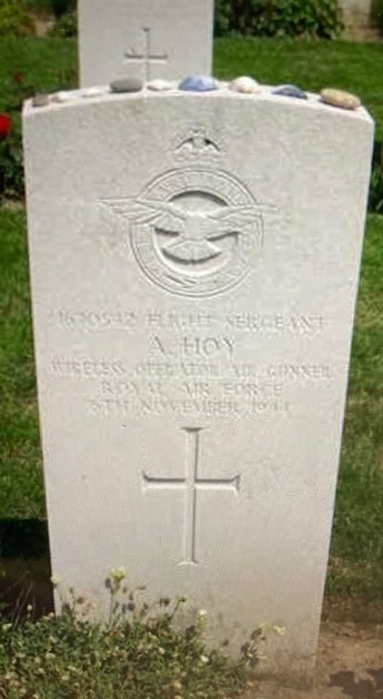 A photo of a British soldier's grave in Germany. A military insignia is embossed on the headstone, as is a Christian cross. Stones sit across the top of the headstone.