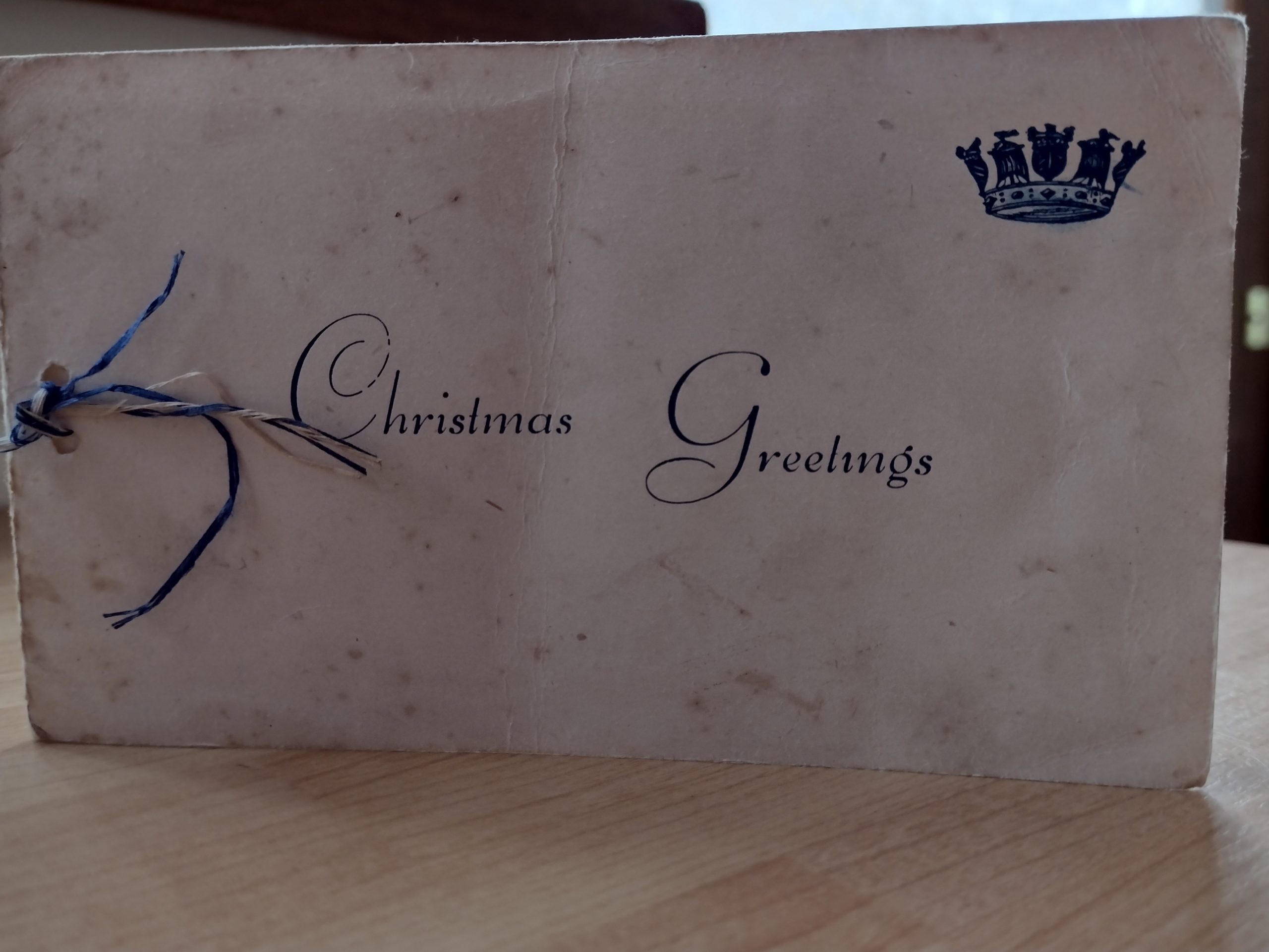 An old Christmas card reads 'Christmas Greetings'. The card is old and cream coloured. Blue and white string binds the card together. In the upper right corner, there is a blue crown. 