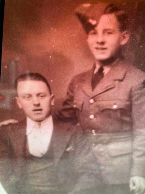 Two men pose for a portrait. An incredibly young looking boy is standing in uniform next to a sitting man in a suit.