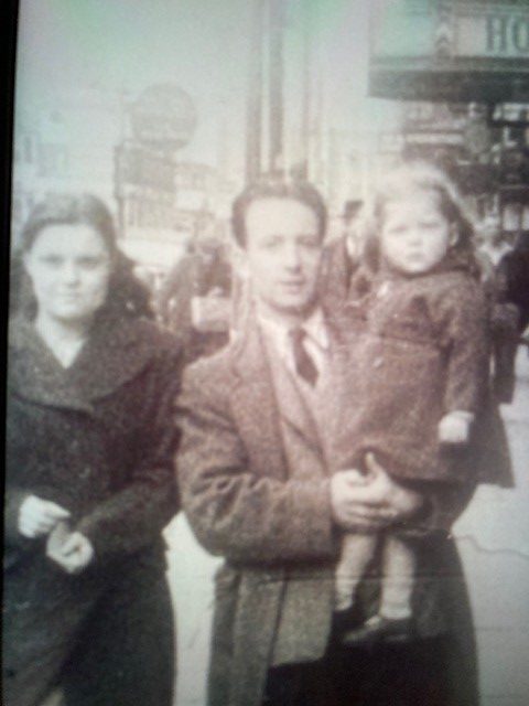 A man holds his child in his arms while his wife stands beside him. Belfast can be seen in the background.