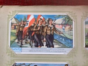 A page of a postcard album depicting life at a summer camp for Hitler Youth children. In the one visible here, girls march with a swastika flag.