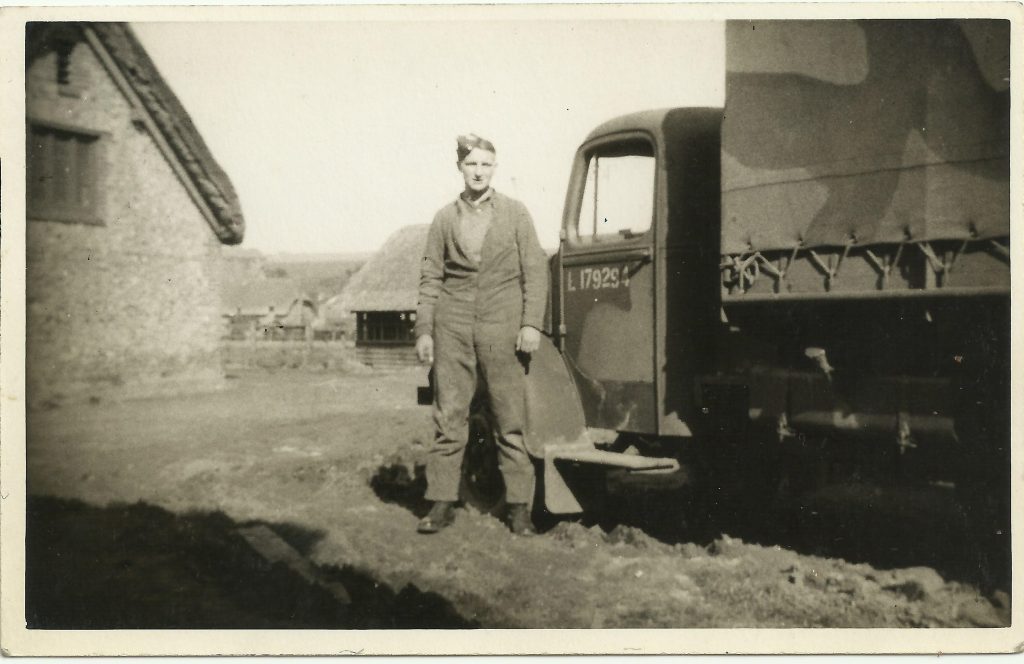 A man in uniform stands next to an armed forces vehicle pained in camouflage. A farmyard is seen behind him.