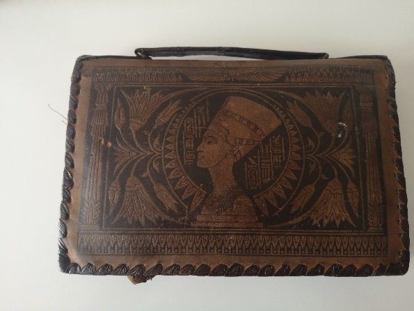 A leather handbag features beautiful and intricate decoration of Egyptian symbols and imagery. The main image is of a woman with a headdress facing left. 