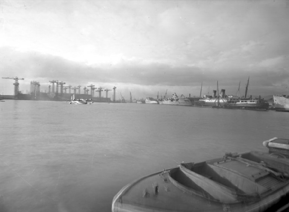 A view over the Musgrave Channel with boats docked to the left and cranes in the far background. A sea plane is coming in to land. 