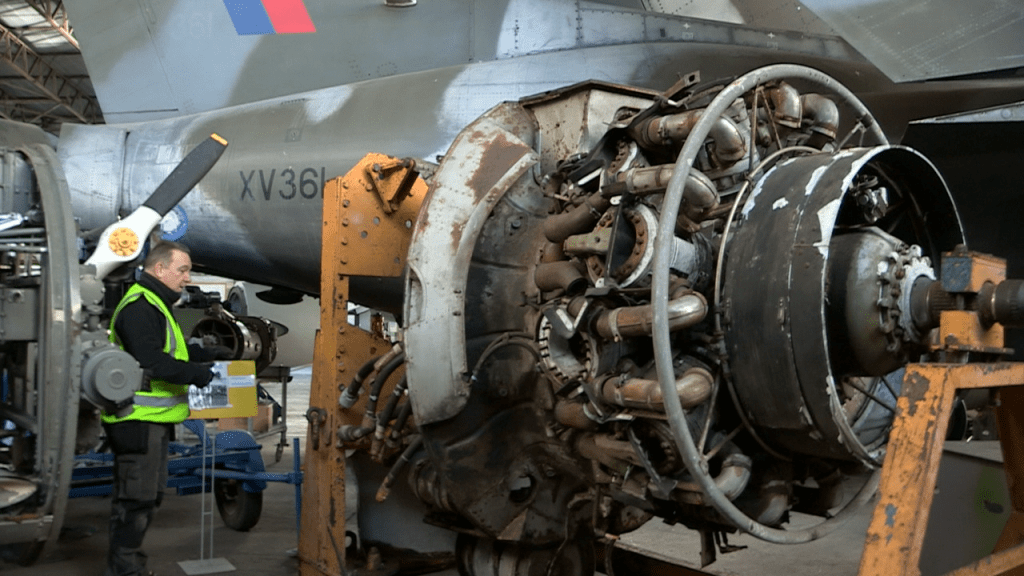 A photo of a large engine. The engine is  a little rusty and old. Beside the engine is a man wearing a hi-vis vest. He is about the same size as the engine. 