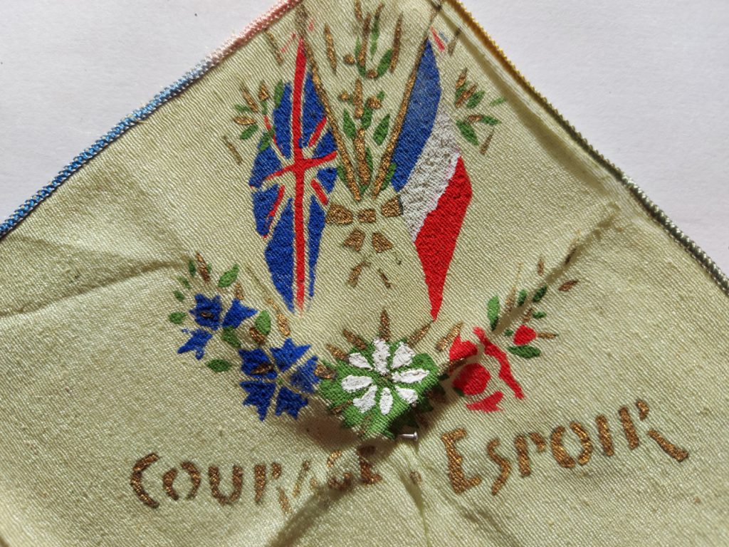 A corner of a handkerchief with beautiful decoration. The French and United Kingdom flag are crossed and surrounded by flowers. At the bottom, there are flowers in white, blue, and red. Under the flowers are the words: 'courage & espoir'.