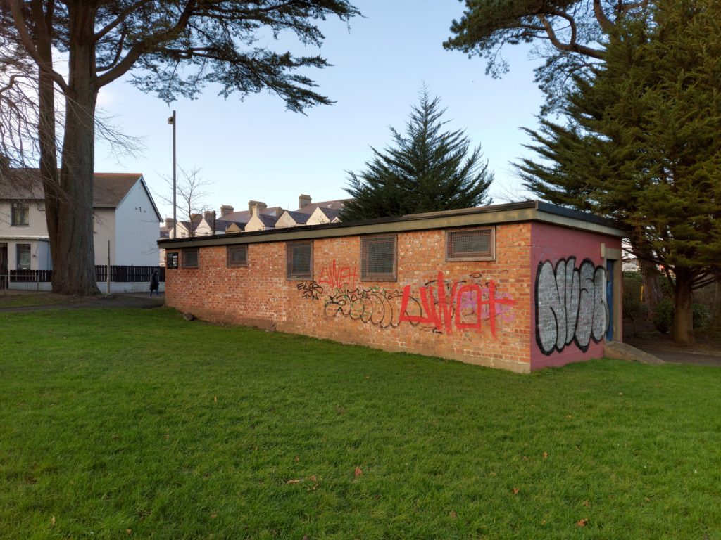 A bomb shelter is seen surrounded by grass. In the background are houses. The shelter has been graffitied. Windows have been installed.